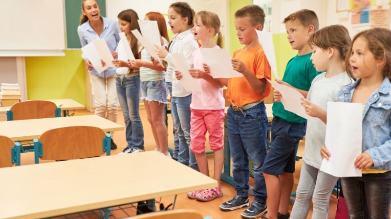 Eine Gruppe Grundschulkinder übt im Klassenzimmer mit einer Musikpädagogin singen